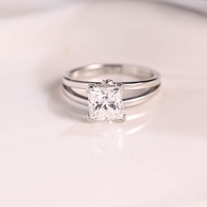 May include: A silver ring with a square-shaped diamond in the center. The ring has a double band design.