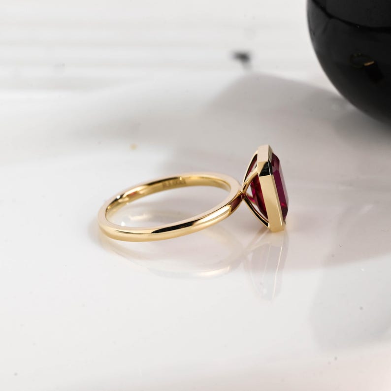 May include: A gold ring with a rectangular, deep red gemstone. The ring band is thin and polished, reflecting light. The gemstone is set in a bezel setting, showcasing its shape and color. The ring is displayed on a white surface.