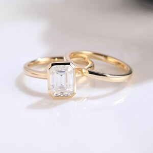 May include: A yellow gold ring set featuring a central emerald-cut clear gemstone held in a bezel setting, accompanied by a plain, thin yellow gold band. Both rings have a polished, smooth finish and a simple, clean design with no additional stones.