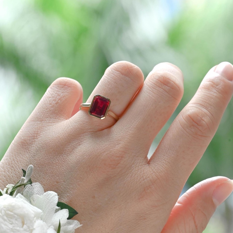 Puede incluir: Un anillo de oro con una piedra preciosa roja de corte esmeralda rectangular montada en una banda de metal sencilla. La piedra es de color rojo intenso y transl&uacute;cida, sujeta por cuatro garras. La banda es de metal dorado liso y pulido.