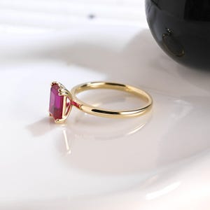 May include: A gold ring featuring a rectangular, emerald-cut vibrant pink gemstone set in a four-prong setting. The band is thin, polished, and circular, showcasing a clean, simple design that highlights the deep color of the central stone.
