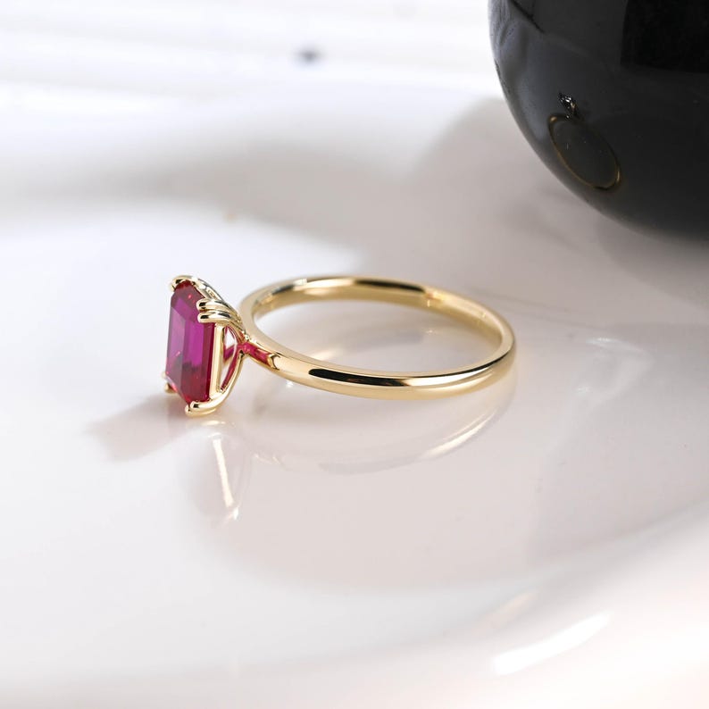 May include: A gold ring featuring a rectangular, emerald-cut vibrant pink gemstone set in a four-prong setting. The band is thin, polished, and circular, showcasing a clean, simple design that highlights the deep color of the central stone.