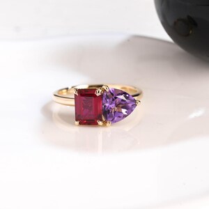 May include: A gold ring featuring a rectangular red gemstone and a triangular purple gemstone. The ring has a simple band and the gemstones are set close together, creating a unique design. The gemstones are vibrant and eye-catching.