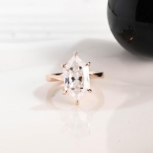 May include: A rose gold engagement ring with a large, clear, hexagonal-cut gemstone. The ring's band is smooth and polished, and the gemstone is set with six prongs. The ring is displayed on a white surface.