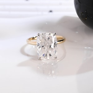 May include: A ring featuring a large, clear, cushion-cut gemstone set in a four-prong setting. The band is made of polished yellow gold, providing a warm contrast to the brilliant, multifaceted white stone that reflects light from its surface.