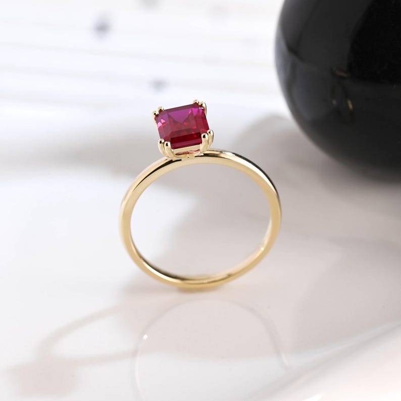 May include: A gold ring featuring a single, square-cut, vibrant red gemstone held in a four-prong setting. The band is thin and polished, showcasing a simple design that highlights the deep crimson color of the faceted stone at the top.