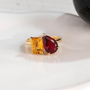 May include: A gold ring with two gemstones: a rectangular yellow stone and a pear-shaped red stone. The ring has a polished gold band. The gemstones are set close together, creating a unique design. The ring is a piece of jewellery.