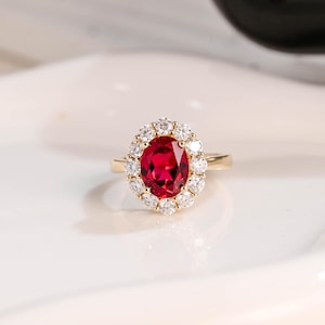 Puede incluir: Un anillo de oro con un gran rubí ovalado rodeado por un halo de diamantes más pequeños.