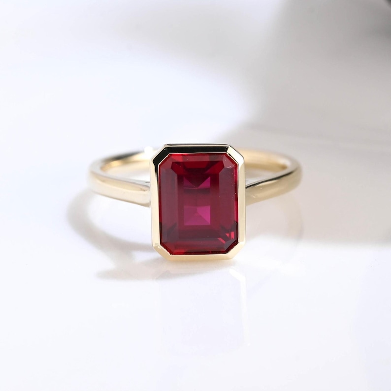 Puede incluir: Un anillo de oro con una piedra preciosa roja de corte esmeralda rectangular montada en un engaste de bisel liso. La banda es de oro pulido y la piedra est&aacute; colocada en el centro, mostrando facetas de color rojo intenso y un dise&ntilde;o geom&eacute;trico.