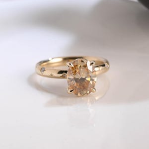 Op de afbeelding: Een gouden ring met een ovale, lichtbruine edelsteen. De gouden band heeft kleine stervormige gravures en kleine heldere stenen. De ring staat tegen een witte achtergrond, waardoor de details en de glans worden benadrukt.