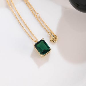 Könnte beinhalten: Eine goldfarbene Halskette mit einem rechteckigen, smaragdgrünen Edelsteinanhänger. Der Anhänger ist in einen goldenen Rahmen gefasst und hängt an einer zarten Goldkette. Die Halskette wird auf einer weißen Oberfläche präsentiert.