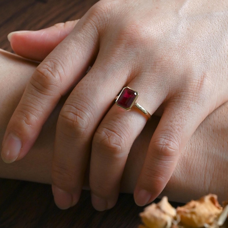 Puede incluir: Un anillo de oro con una piedra preciosa rectangular de color rojo intenso en un engaste de bisel liso. La banda es fina y pulida, mostrando un dise&ntilde;o que resalta el color vibrante y la forma geom&eacute;trica de la piedra central.