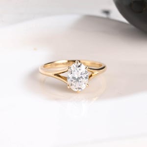 Könnte beinhalten: Ein goldener Ring mit einem großen, ovalförmigen, klaren Edelstein. Das Band des Rings teilt sich auf beiden Seiten des Edelsteins in ein zartes Design auf, wodurch ein eleganter und moderner Look entsteht. Der Ring ist vor einem weißen Hintergrund platziert.