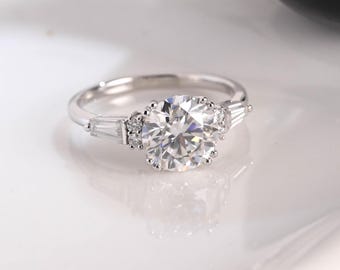 Può includere: Un anello color argento con una grande pietra centrale rotonda a taglio brillante. La pietra centrale è affiancata da due pietre rettangolari a taglio baguette su ciascun lato, con minuscole pietre rotonde incastonate tra esse.