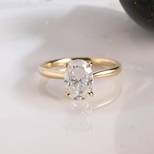 May include: A gold ring featuring a single oval-cut clear gemstone held in a four-prong setting. The band is smooth and polished, showcasing a classic design with a tapered shank that leads up to the central stone, reflecting light clearly.