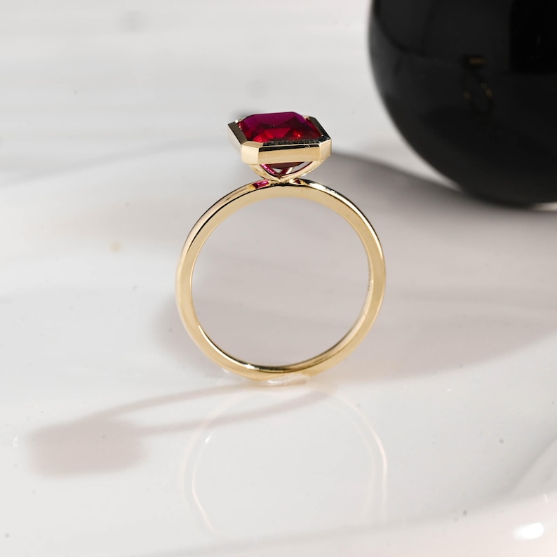 May include: A gold ring with a large, rectangular, deep red gemstone. The ring's band is thin and polished, and the gemstone is set in a gold bezel. The ring is displayed on a white surface.