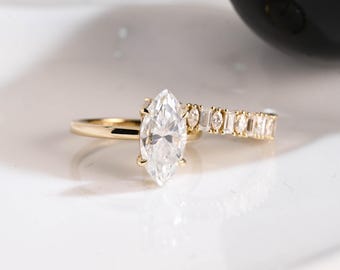 May include: Two gold rings with clear, sparkling gemstones. One ring features a marquise-cut stone, while the other has a row of smaller, rectangular and round stones. The rings are set against a white background.