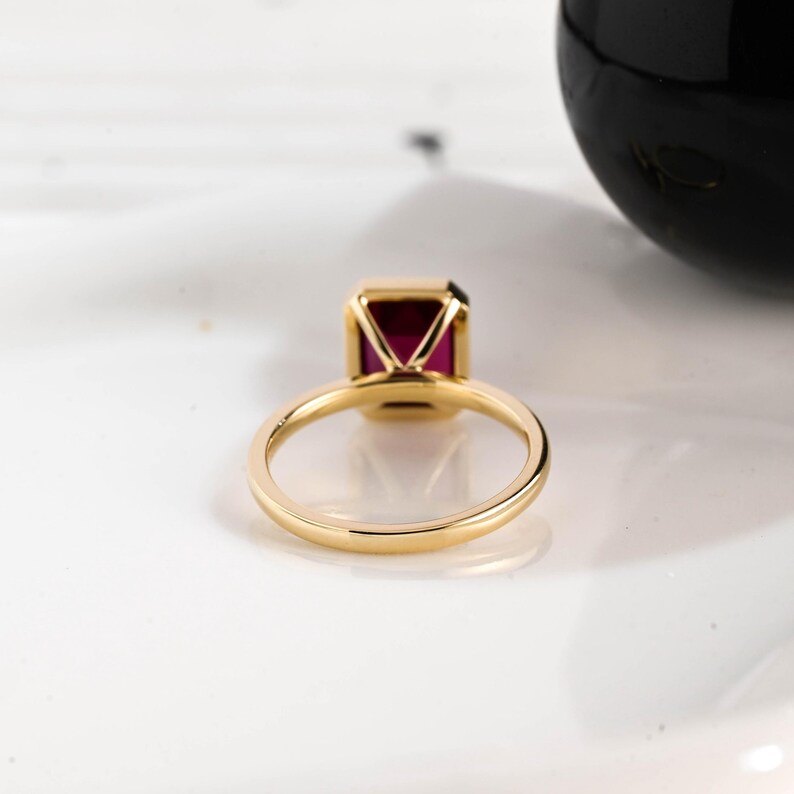May include: A gold ring with a rectangular, deep red gemstone. The ring's band is a simple, polished gold, and the gemstone is set in a gold bezel. The ring is displayed on a white surface.