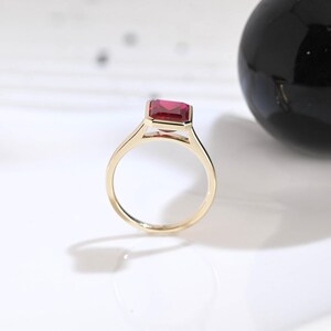Puede incluir: Un anillo de oro con una piedra preciosa rectangular, facetada y de color rojo intenso, montada en un engaste de bisel. La banda es fina y pulida, mostrando un dise&ntilde;o limpio que resalta el color vibrante y la claridad de la piedra central.
