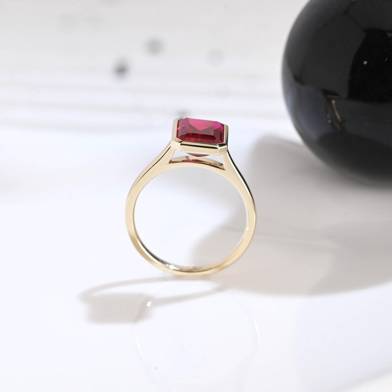 Puede incluir: Un anillo de oro con una piedra preciosa rectangular, facetada y de color rojo intenso, montada en un engaste de bisel. La banda es fina y pulida, mostrando un dise&ntilde;o limpio que resalta el color vibrante y la claridad de la piedra central.