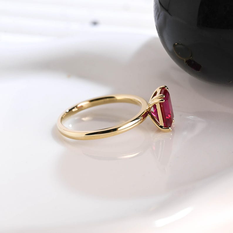 May include: A gold ring featuring a rectangular, faceted, deep red gemstone set in a four-prong setting. The band is smooth and polished, showcasing a classic design that highlights the vibrant color and clarity of the central stone.