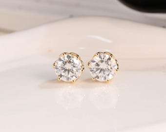 May include: A pair of gold-coloured stud earrings featuring large, clear, round, faceted stones. The earrings are set in a six-prong setting, and the stones sparkle. The earrings are displayed on a white surface.
