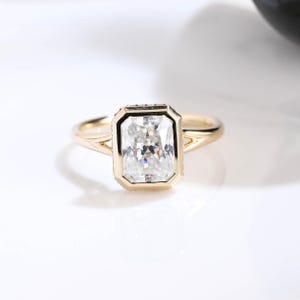 Könnte beinhalten: Ein Goldring mit einem rechteckigen, im Radiant-Schliff geschliffenen, klaren Edelstein in einer Zargenfassung. Die Ringschiene hat ein geteiltes Design, das sich zum Mittelstein hin verjüngt und für ein poliertes, reflektierendes Aussehen sorgt.