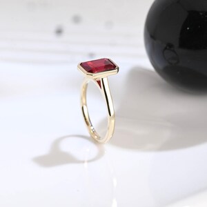 Puede incluir: Un anillo de oro con una piedra preciosa roja rectangular de corte esmeralda engastada en una banda de metal pulido. El engaste sujeta firmemente la piedra roja vibrante, mostrando un dise&ntilde;o limpio con un acabado liso y brillante.