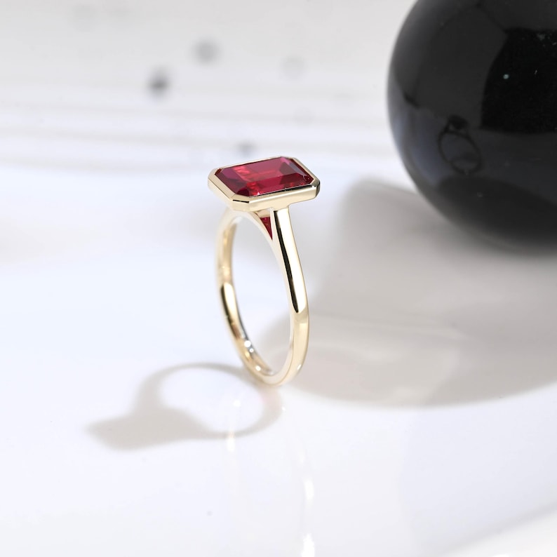 Puede incluir: Un anillo de oro con una piedra preciosa roja rectangular de corte esmeralda engastada en una banda de metal pulido. El engaste sujeta firmemente la piedra roja vibrante, mostrando un dise&ntilde;o limpio con un acabado liso y brillante.