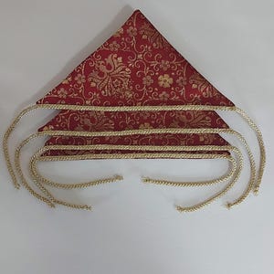 May include: A red and gold decorative textile with a triangular shape. The fabric features a gold floral pattern. Gold braided trim and gold cords extend from the textile, creating a decorative design.