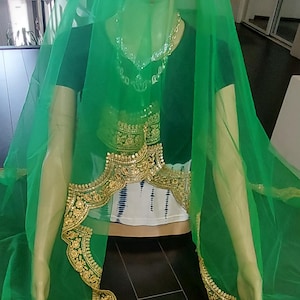 May include: A mannequin draped in a green sheer veil and a black top. The veil features a gold embroidered border. A silver necklace and a gold embroidered belt are also visible. The mannequin stands on a dark wood floor.