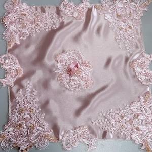 Puede incluir: Un cuadrado de satén rosa pálido con intrincados encajes florales y adornos de cuentas. Las esquinas y el centro presentan diseños de encaje detallados. Una pequeña cinta rosa está unida al encaje central. El tema general es delicado y femenino.