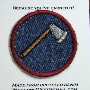May include: A circular merit badge with a denim background and a red border. The badge features an embroidered axe with a silver head and a brown handle. The text "MERIT BADGE" and "BECAUSE YOU'VE EARNED IT!" are at the top.