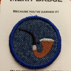 May include: A blue denim merit badge with a brown pipe design. The badge is round and has a blue border. The text "MERIT BADGE" is printed in black at the top of the card. The text "BECAUSE YOU'VE EARNED IT!" is printed in black below the badge. The text "MADE FROM UPCYCLED DENIM" is printed in black at the bottom of the card. The text "WAGGONSWEST@GMAIL.COM" is printed in black below the text "MADE FROM UPCYCLED DENIM". The text "WAGGONSWEST.ETSY.COM" is printed in black below the text "WAGGONSWEST@GMAIL.COM".