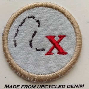May include: Round patch with a light blue center, embroidered with a black design and a red "X". The patch has a tan border and the text "MADE FROM UPCYCLED DENIM" at the bottom. This patch is likely for clothing or accessories.
