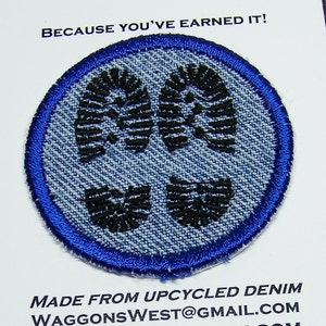 May include: Blue denim patch with three black footprints, surrounded by a blue border. The text "Because you've earned it!" is printed above the patch. The text "Made from upcycled denim" is printed below the patch. The text "WaggonsWest@gmail.com" is printed below the text "Made from upcycled denim".