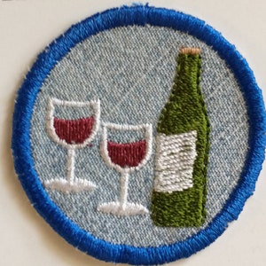 May include: Embroidered patch featuring a wine bottle and two wine glasses filled with red wine. The design is set against a light blue background with a blue border. The patch is round and appears to be made of fabric.
