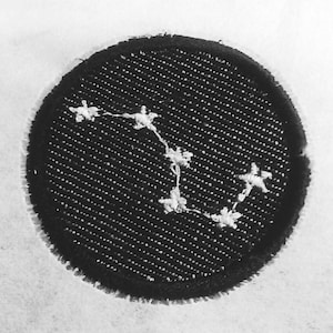 May include: A circular black embroidered patch featuring a white stitched constellation pattern. The design consists of six small white star-shaped clusters connected by thin white lines, all set against a textured black fabric background.