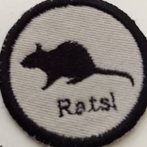 May include: A circular patch with a black rat silhouette on a light gray background. The word "Rats!" is embroidered in black below the rat. The patch has a black border.