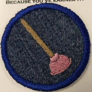 May include: Embroidered patch featuring a pink plunger with a tan handle, set against a denim-like background. The patch is circular, with a blue border. The text "BECAUSE YOU'VE EARNED IT!" is visible at the top.