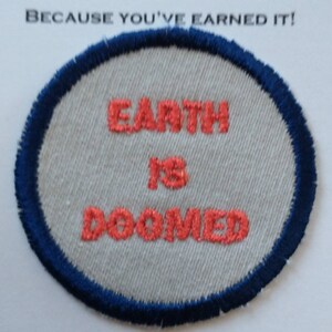 May include: A white oval patch with a blue border. The patch has red embroidered text that reads "Earth is Doomed".