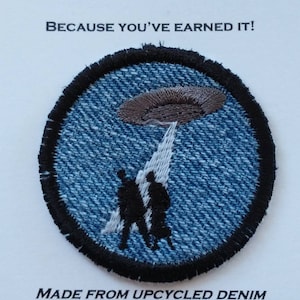 May include: A blue denim patch with a black embroidered border. The patch features a gray UFO with a white beam of light shining down on three black silhouettes of people. The text "MERIT BADGE" is printed above the patch and "BECAUSE YOU'VE EARNED IT!" is printed below the patch. The text "MADE FROM UPCYCLED DENIM" is printed below the patch, along with the email address "WAGGONSWEST@GMAIL.COM" and the website address "WAGGONSWEST.ETSY.COM".