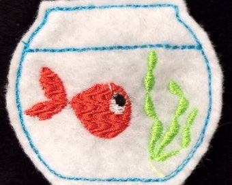 May include: A white felt patch shaped like a fishbowl with blue embroidered outlines. Inside the bowl, there is a bright orange fish with a black eye and a green embroidered aquatic plant. The design is stitched onto a white felt base.