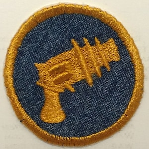 May include: A gold embroidered patch with a blue denim background. The patch features a stylized image of a futuristic ray gun.