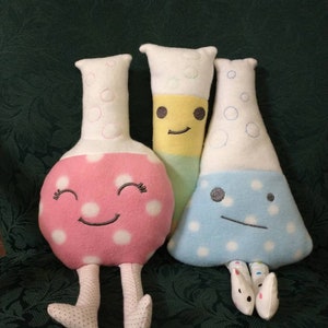 May include: Three plush toys shaped like beakers with embroidered faces. The beaker on the left is pink with white polka dots, the middle beaker is yellow with green stripes, and the beaker on the right is blue with white polka dots.