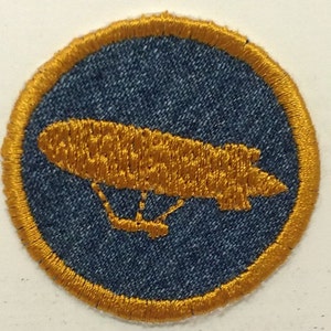 May include: A blue denim patch with a gold embroidered outline and a gold embroidered image of an airship.