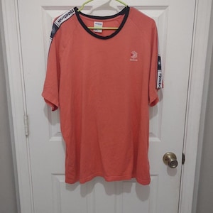 Op de afbeelding: Koraalrood T-shirt met een zwart-witte bies rond de kraag en mouwen. Het shirt heeft het Reebok-logo op de borst en de naam Reebok op de mouwen.