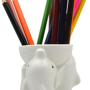 Peut inclure: Un porte-crayons en céramique blanche en forme de deux dauphins, contenant une collection de crayons de couleur. Les crayons sont de différentes couleurs, dont le noir, l'orange, le vert, le bleu et le rose.
