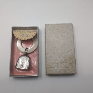 May include: A vintage baby rattle by Webster, presented in its original box. The rattle features a white teething ring and a silver-toned bell charm. The box is pink inside with the text "A Tiny Toy Treasure".