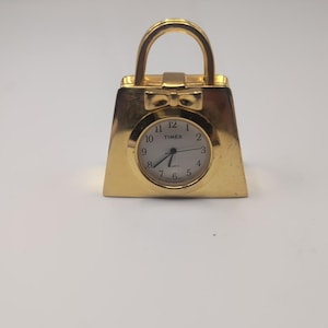 Może przedstawiać: Złoty zegar kwarcowy Timex w kształcie torebki. Tarcza zegara jest okrągła z czarnymi cyframi i wskazówkami. Projekt torebki obejmuje uchwyt i kokardę. Zegar jest na białym tle.